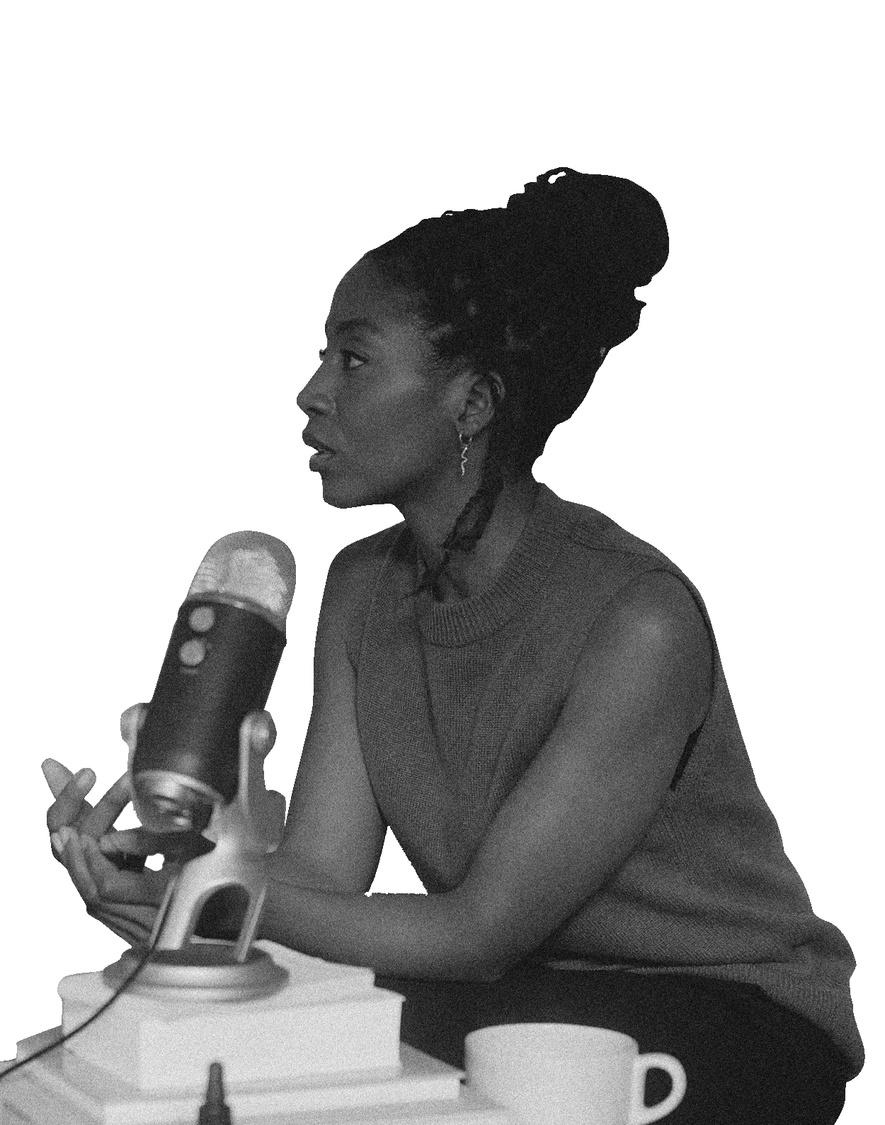 Black and white photo of a woman speaking into a microphone at a desk with books and a coffee mug.