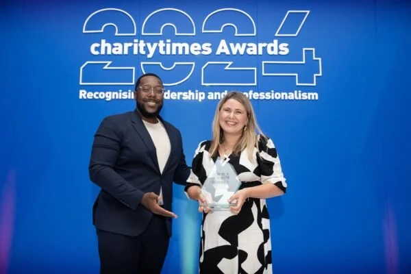 A man and a woman stand together, smiling, during the charitytimes Awards 2024 event, with the woman holding an award. The backdrop features the event name and slogan.