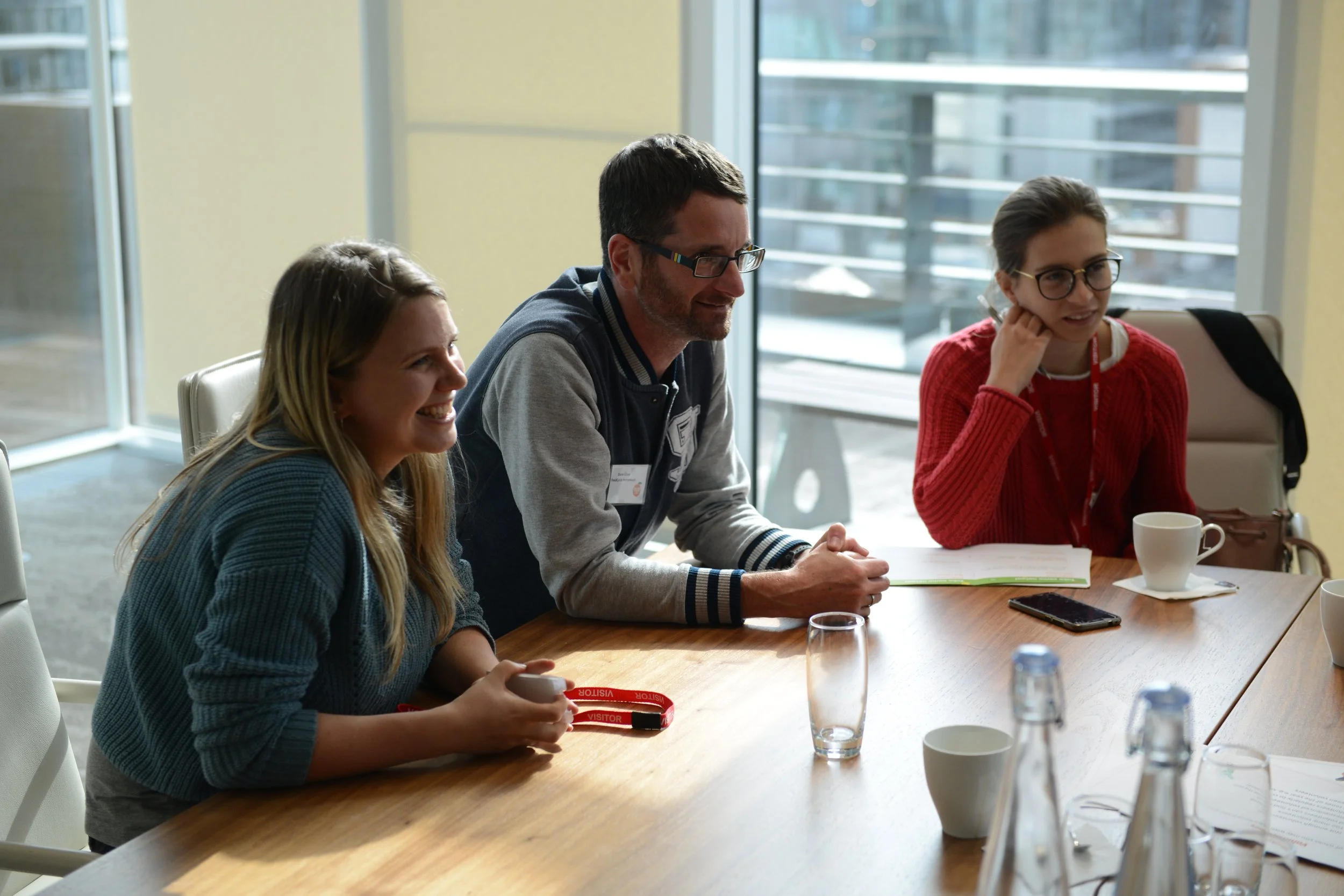 Three people seated at a conference table during a meeting, smiling and engaging in conversation, with coffee cups, glasses, bottles, and notebooks on the table, outside view through large windows.