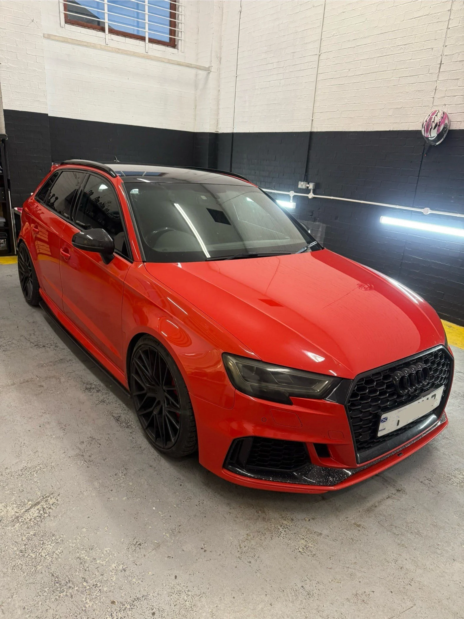 Red Audi sedan with black rims parked inside a garage with white brick walls and black-accented wall with fluorescent lighting.