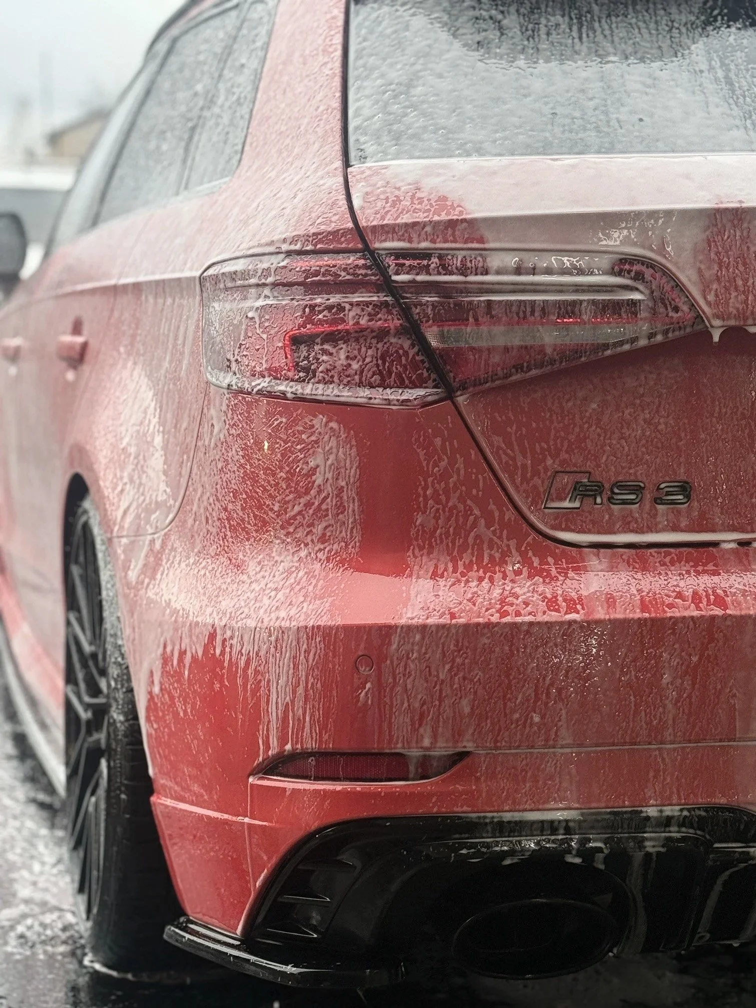 Red Audi RS 3 with water and soap suds on the exterior, parked on a wet surface during rainy weather.