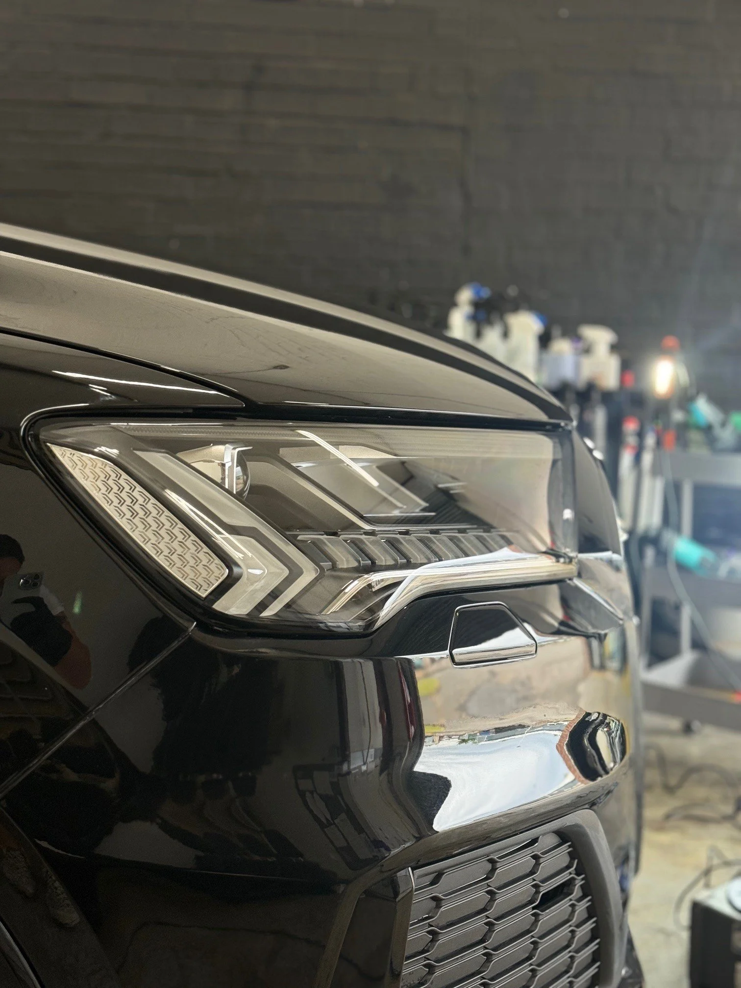 Close-up of the front left side of a black car, showing the headlight, part of the grille, and the hood. The background shows a dark wall and a table with various bottles and tools, indicating a garage or workshop setting.