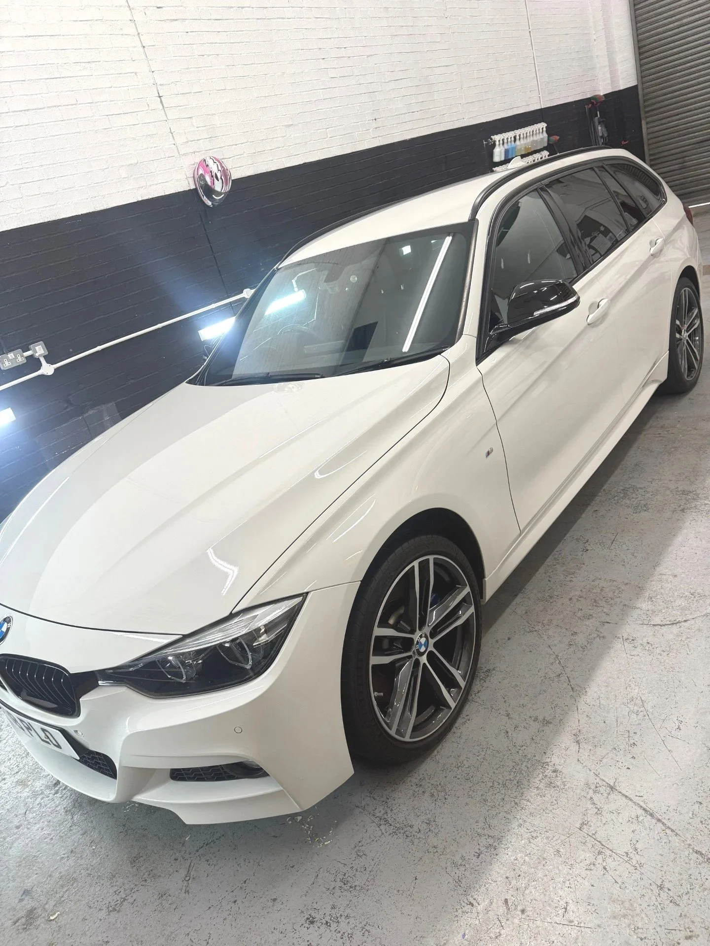 White BMW sedan inside a garage with black and white walls, LED lights, and shelves with cleaning supplies.