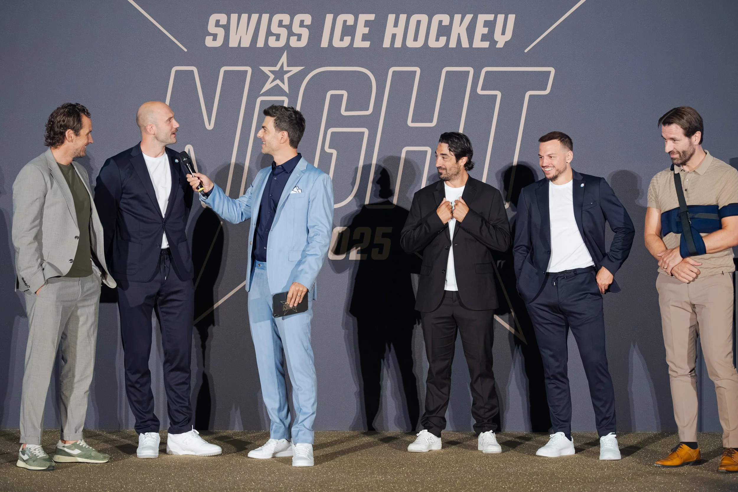 Sieben Männer stehen bei einer Veranstaltung mit dem Titel "Swiss Ice Hockey Night". Ein Mann im grauen Anzug spricht mit einem anderen Mann in einem dunkelblauen Anzug, während die restlichen Männer zuhören und lächeln.