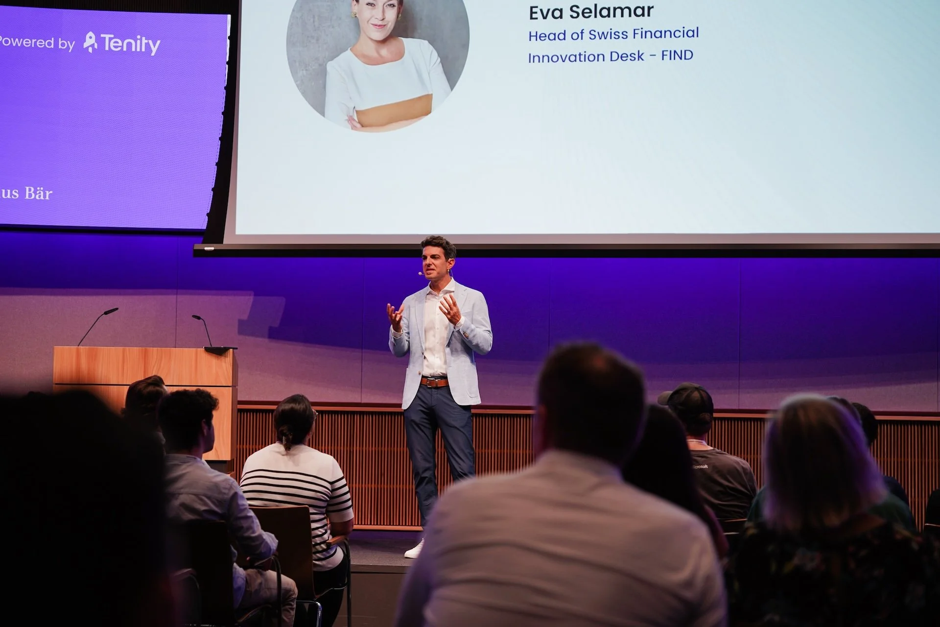 Ein Mann hält eine Präsentation vor einer Gruppe von Zuhörern im Auditorium. Auf der großen Leinwand im Hintergrund sind Fotos und Text zu sehen, inklusive eines Profils von Eva Selamar, Head of Swiss Financial Innovation Desk - FIND.