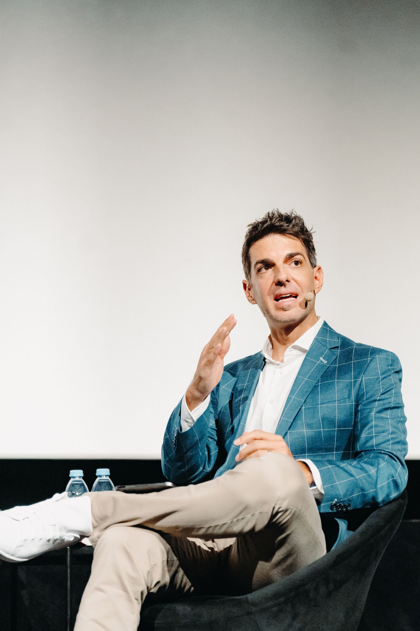 Mann in blauer Anzugjacke und weißen Hemd sitzt in Stuhl und spricht bei einer Präsentation, im Hintergrund eine leere Leinwand.
