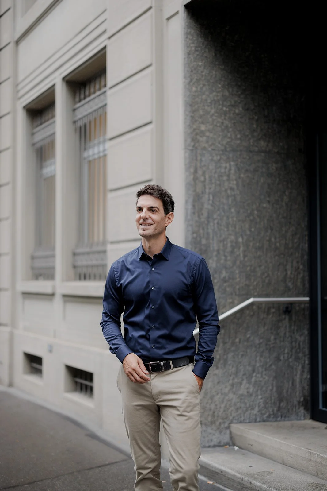Junge Mann in dunklem Hemd und hellen Hosen steht vor einer Gebäudefassade auf der Straße.