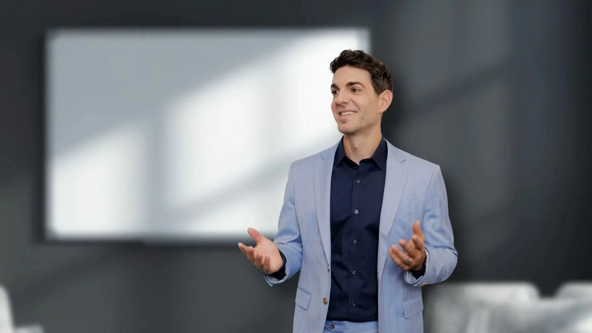 Ein Mann in einem hellblauen Anzug und dunklem Hemd spricht bei einer Präsentation oder einem Meeting in einem modernen Raum, im Hintergrund eine weiße Tafel oder Bildschirm.