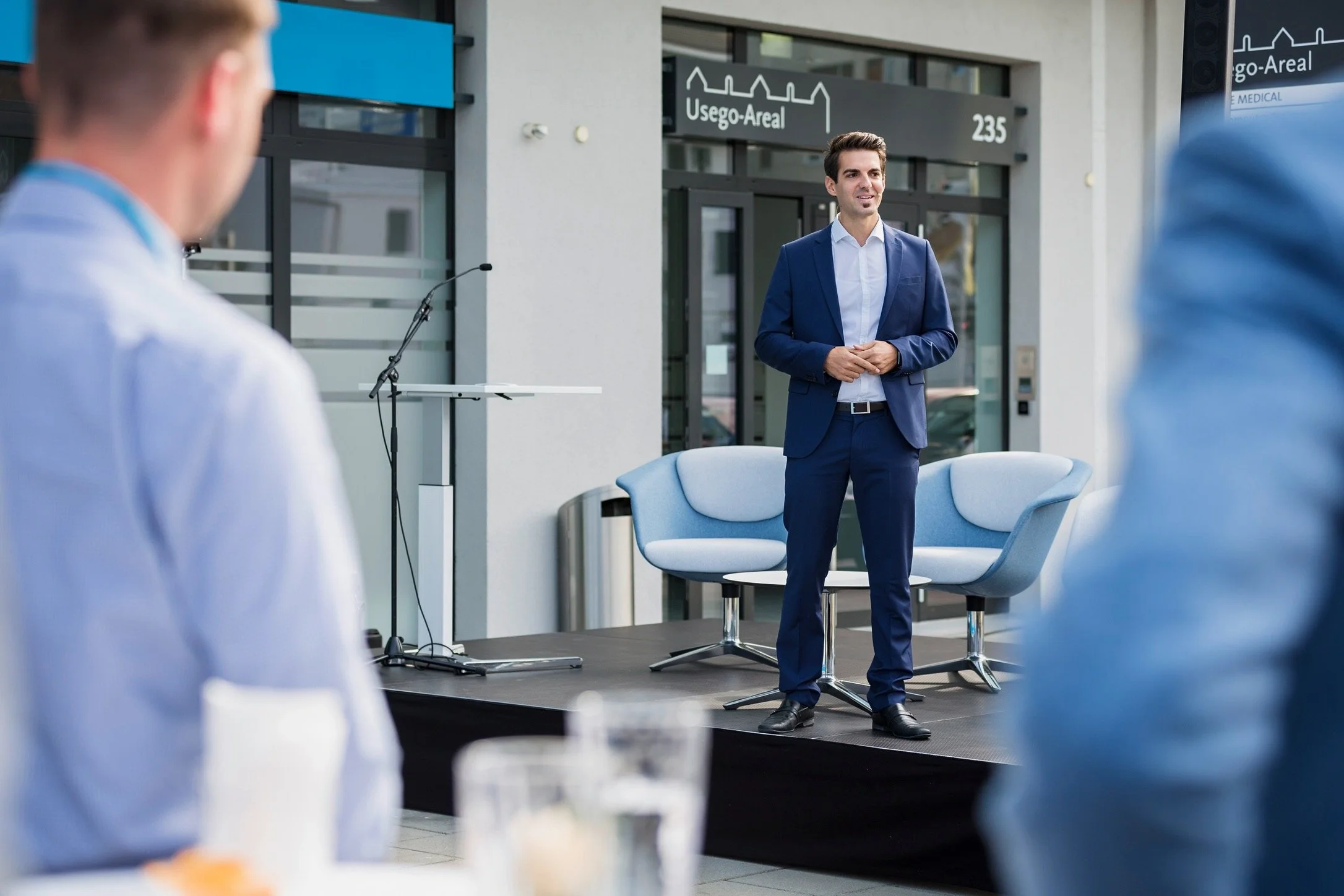 Ein Mann in einem blauen Anzug hält eine Rede bei einer Veranstaltung im Freien, im Hintergrund ist eine Bühne mit einem Mikrofon und Stühlen.