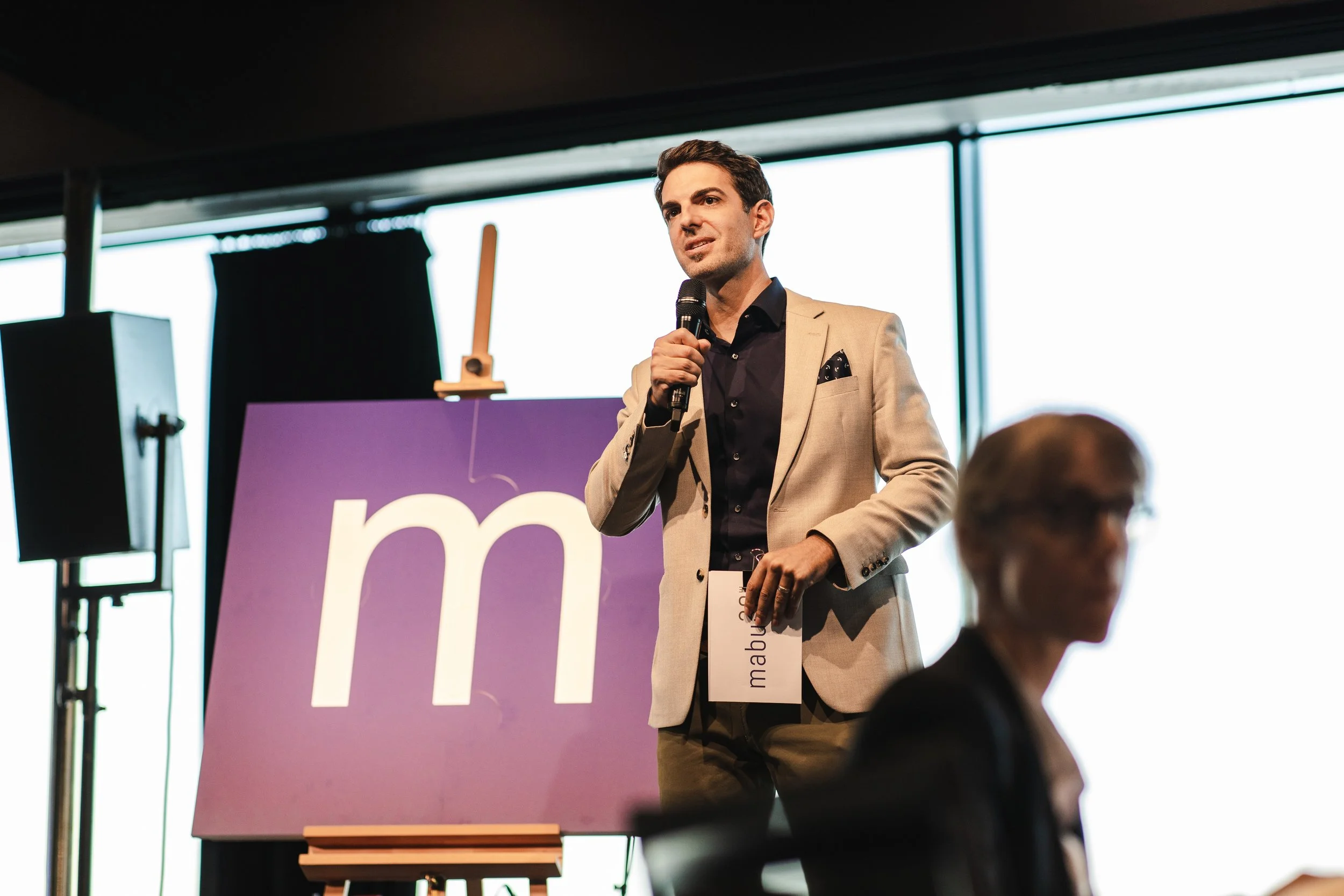 Ein Mann in einem beigefarbenen Anzug hält ein Mikrofon und spricht auf einer Bühne bei einer Veranstaltung. Im Hintergrund ein großes Plakat mit einem weißen M auf lila Hintergrund.