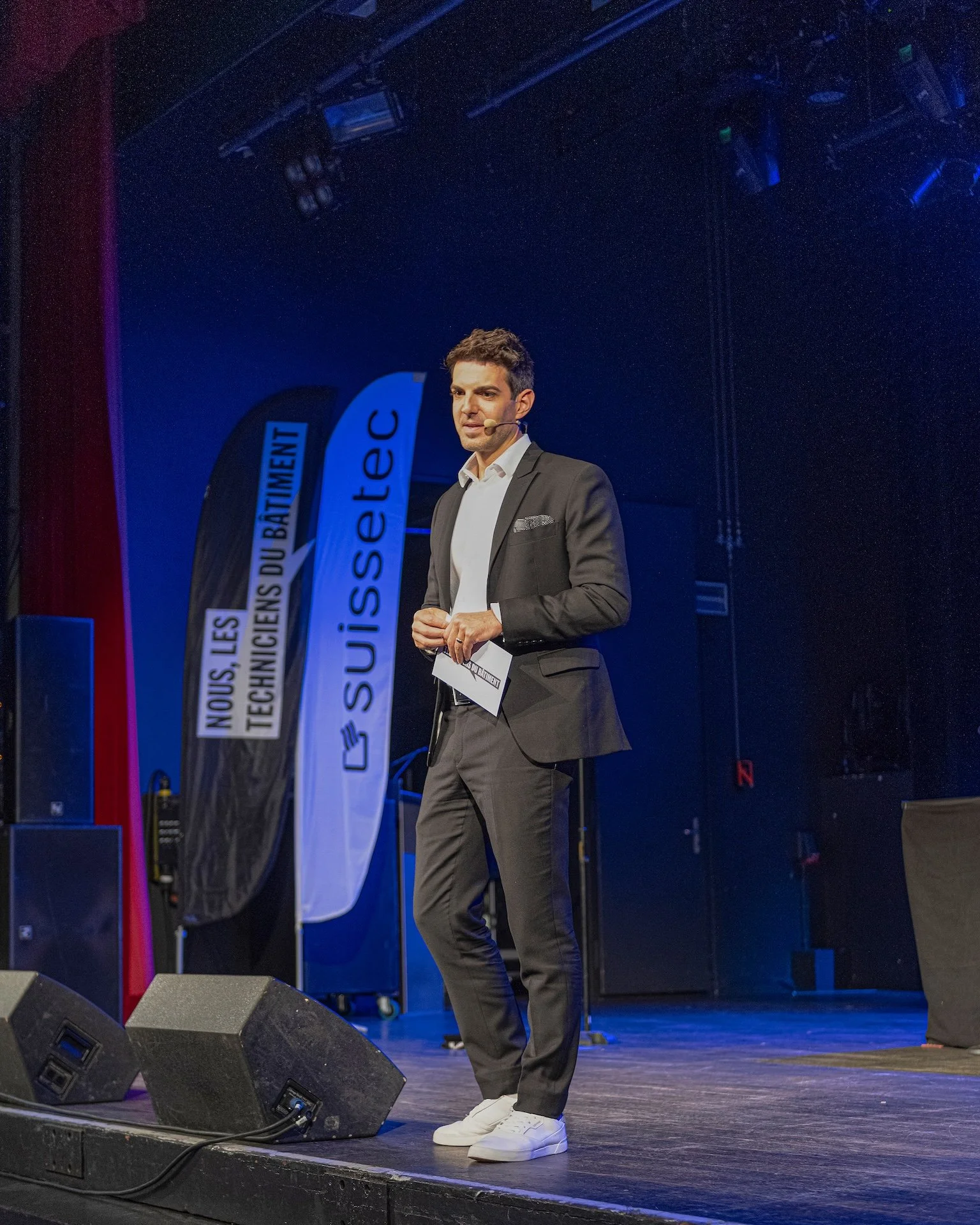 Ein junger Mann in Anzug und Turnschuhen steht auf einer Bühne und hält Notizen in der Hand. Hinter ihm sind Banner mit französischem Text und Lautsprecher, im Hintergrund blaue Beleuchtung.