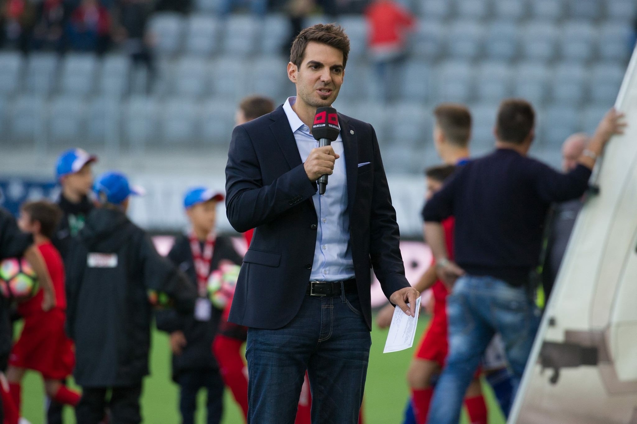 Ein Mann in einem dunklen Anzug hält ein Mikrofon und spricht vor einer Gruppe von Kindern und Erwachsenen auf einem Fußballfeld.