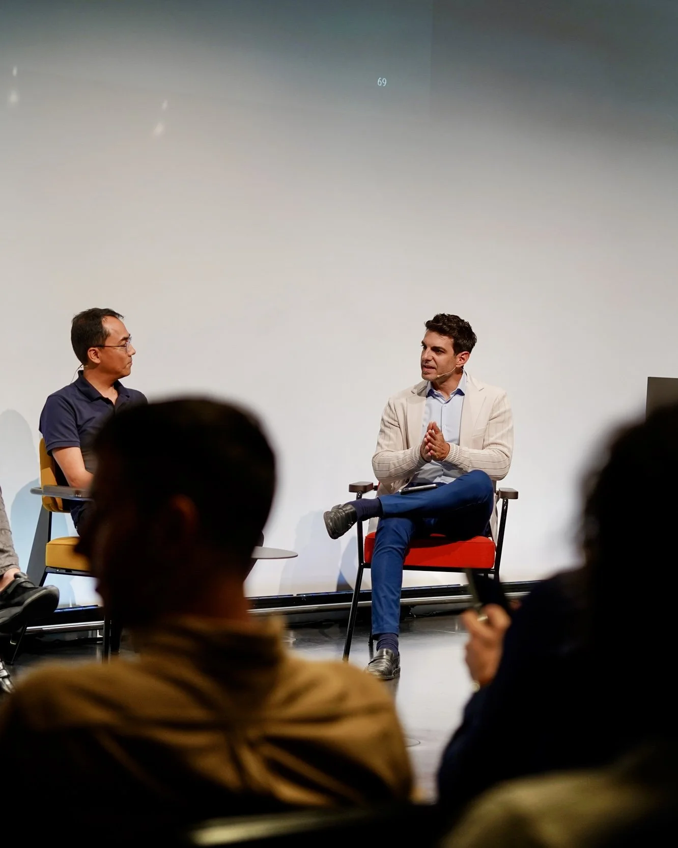 Zwei Männer im Gespräch auf einer Bühne während einer Diskussion, im Vordergrund sitzen mehrere Zuschauer, der Hintergrund ist eine leere weiße Wand.