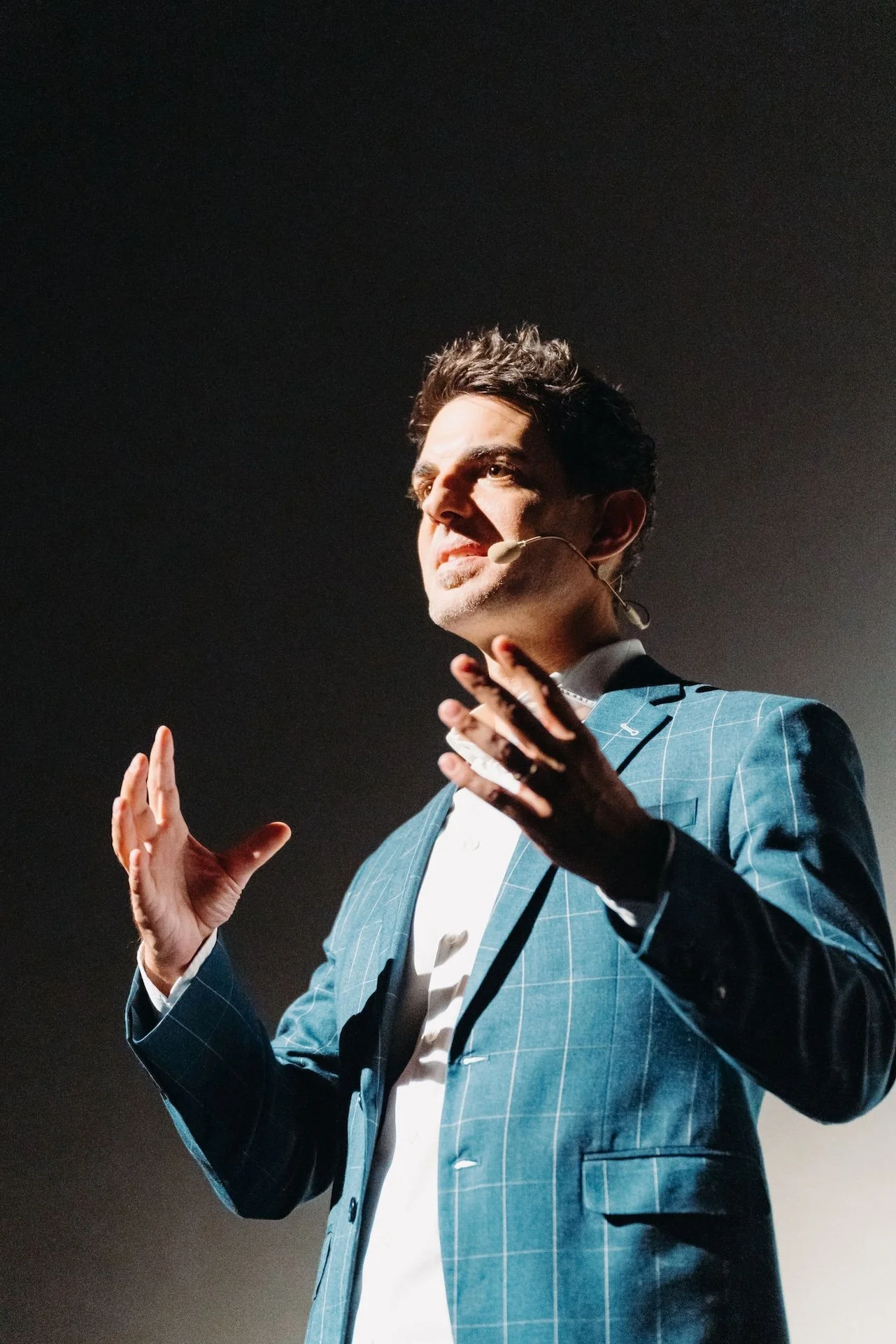 Mann in einem blauen karierten Anzug spricht vor Publikum, trägt ein Headset und macht Gesten mit den Händen.