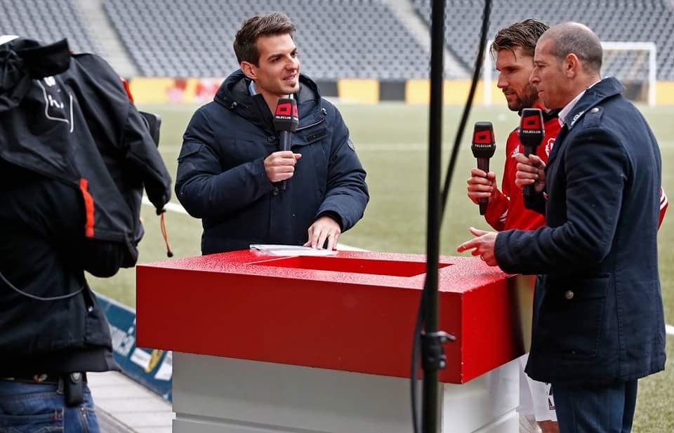 Drei Männer interviewen einen Spieler umgeben von einem Kameramann, vor einem Fußballfeld.