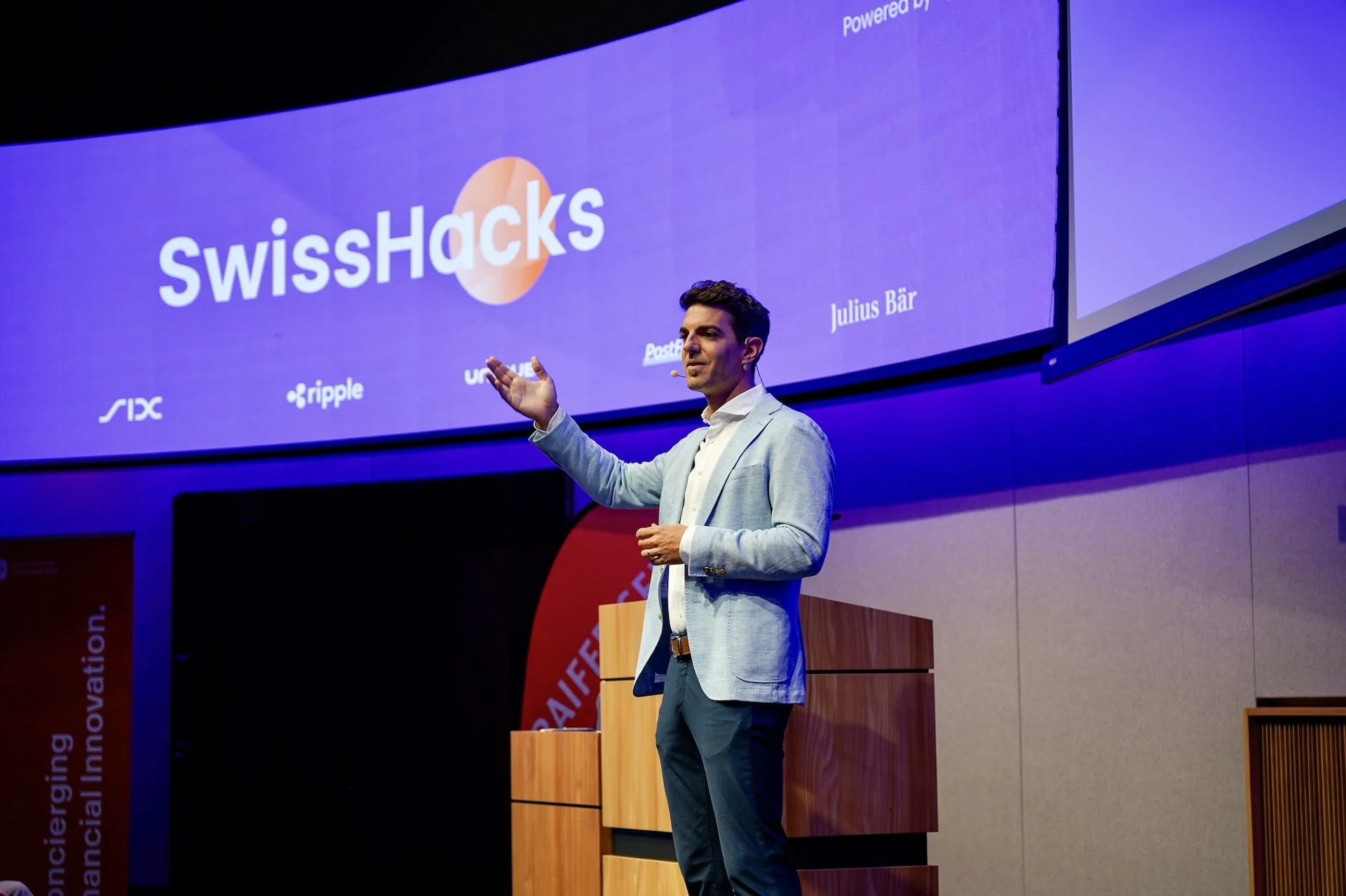 Mann in hellblauem Anzug hält eine Präsentation auf einer Bühne vor einem großen Bildschirm mit dem Logo SwissHacks und weiteren Logos, im Hintergrund ein rotes Panel mit weißen Texten.