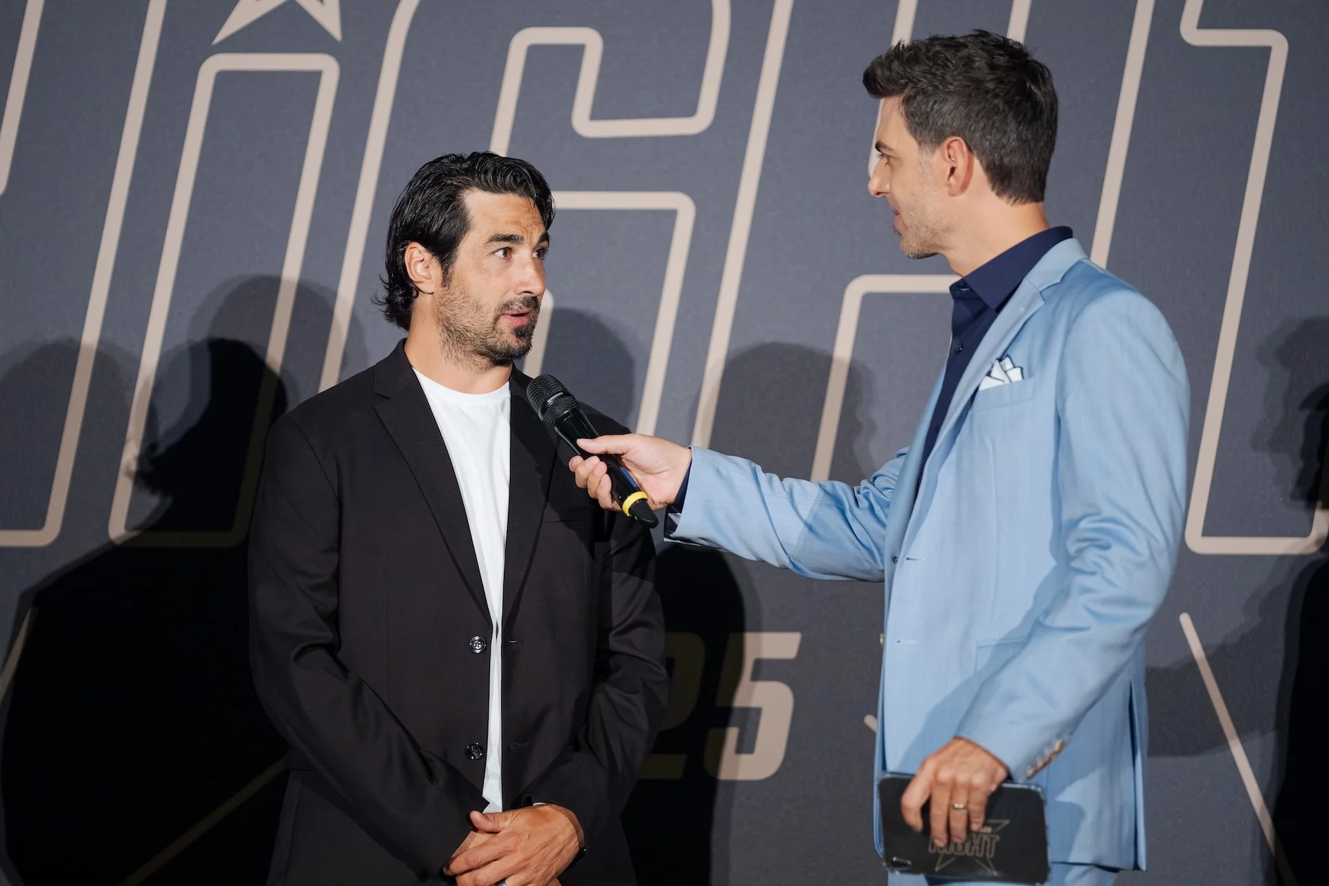 Zwei Männer im Gespräch, einer hält ein Mikrofon, im Hintergrund eine Wand mit geometrischem Muster und Zahlen.