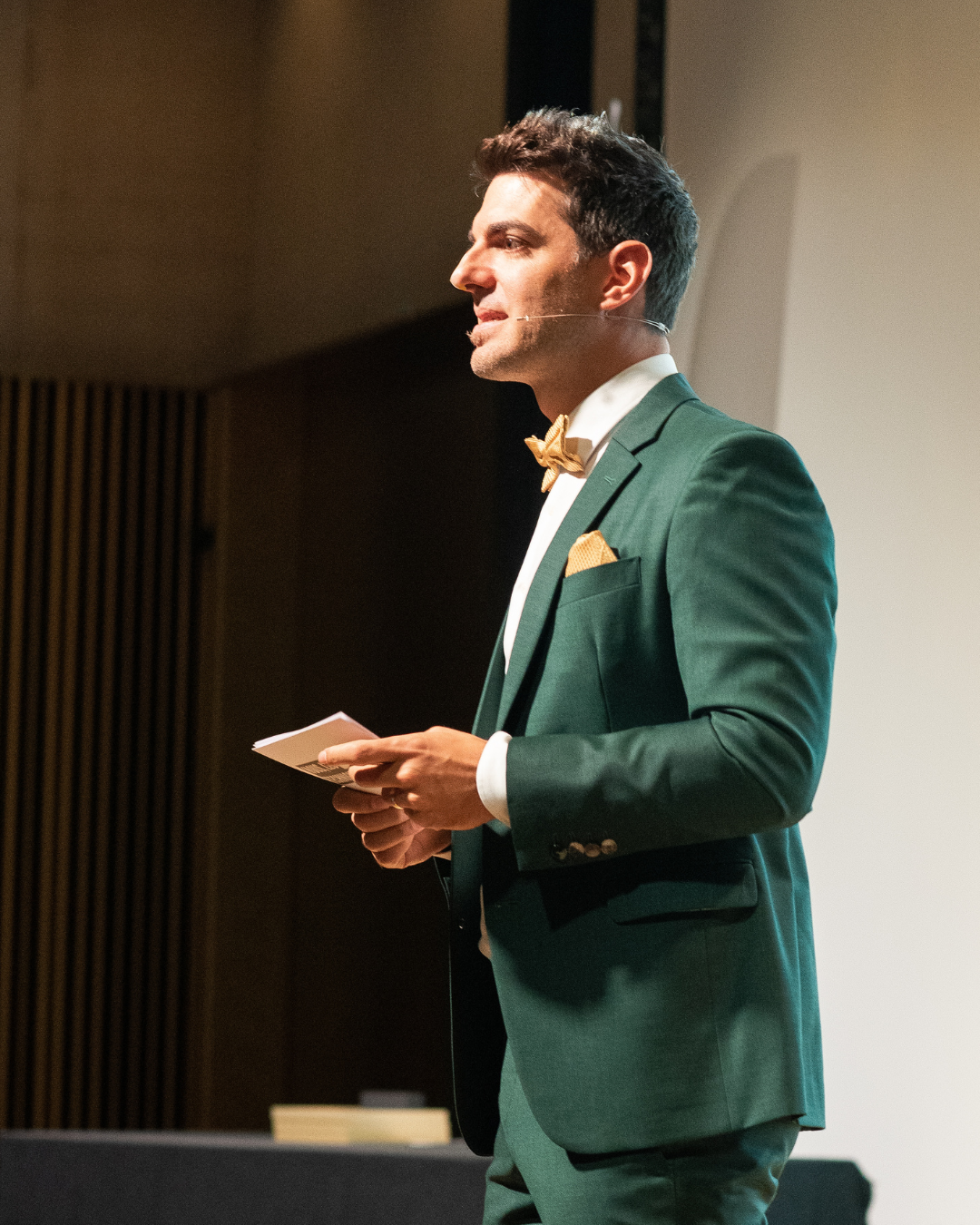 Mann spricht auf einer Bühne bei einer Veranstaltung, trägt einen dunklen Anzug mit goldener Fliege und hält Notizen in den Händen.