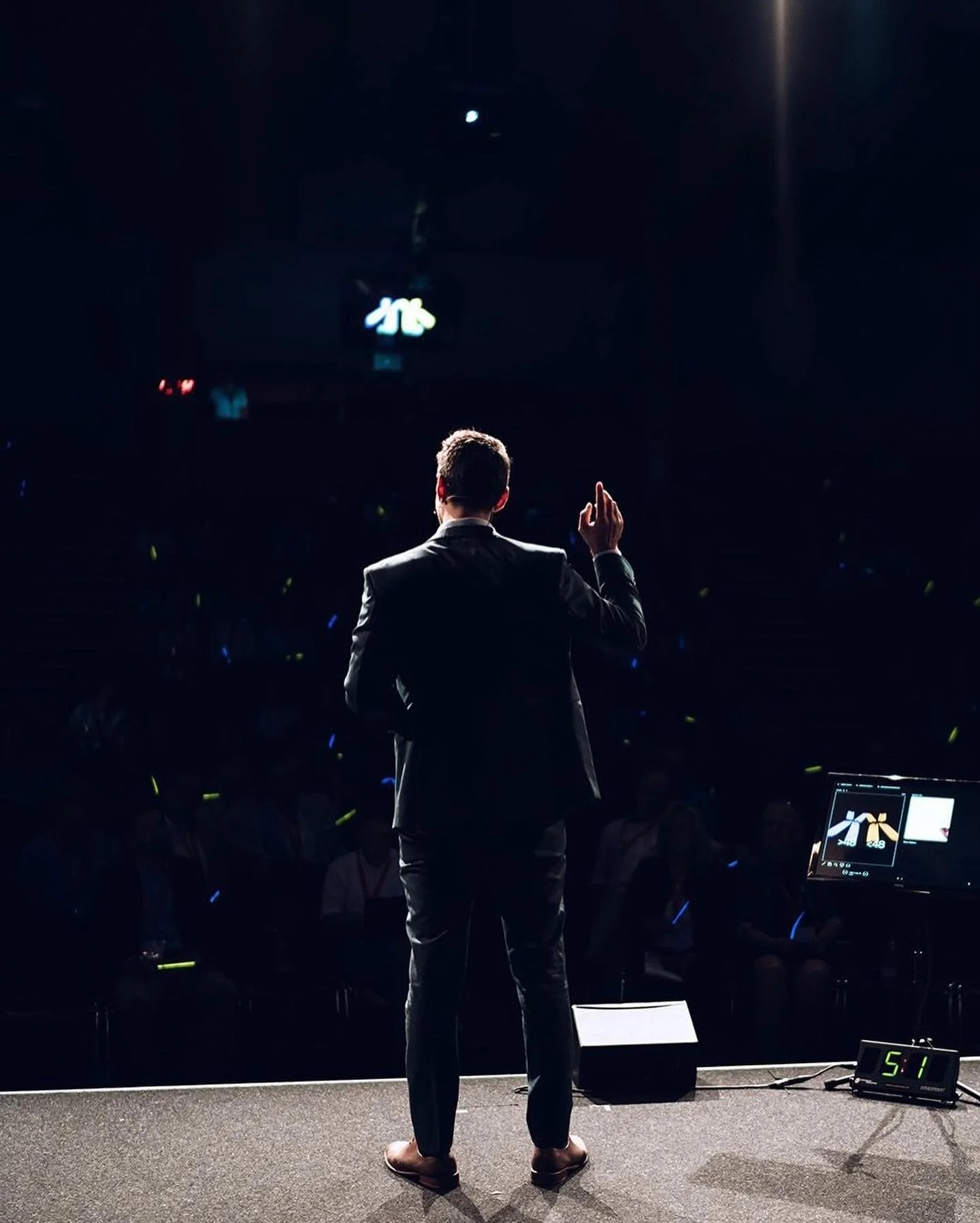 Ein Mann in einem schwarzen Anzug steht auf einer Bühne, von hinten gesehen, während er einem Publikum spricht oder präsentiert. Im Hintergrund ist eine dunkle Atmosphäre mit einigen Lichtakzenten, und auf einem Bildschirm ist ein Logo oder ein Bild sichtbar.