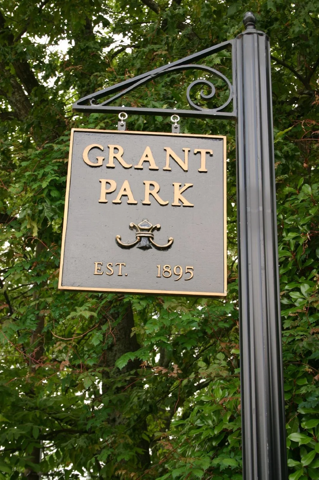 A sign for Grant Park hanging from a decorative black metal pole, with green trees in the background. The sign reads "Grant Park," established in 1895, with a small wrought iron emblem beneath the name.