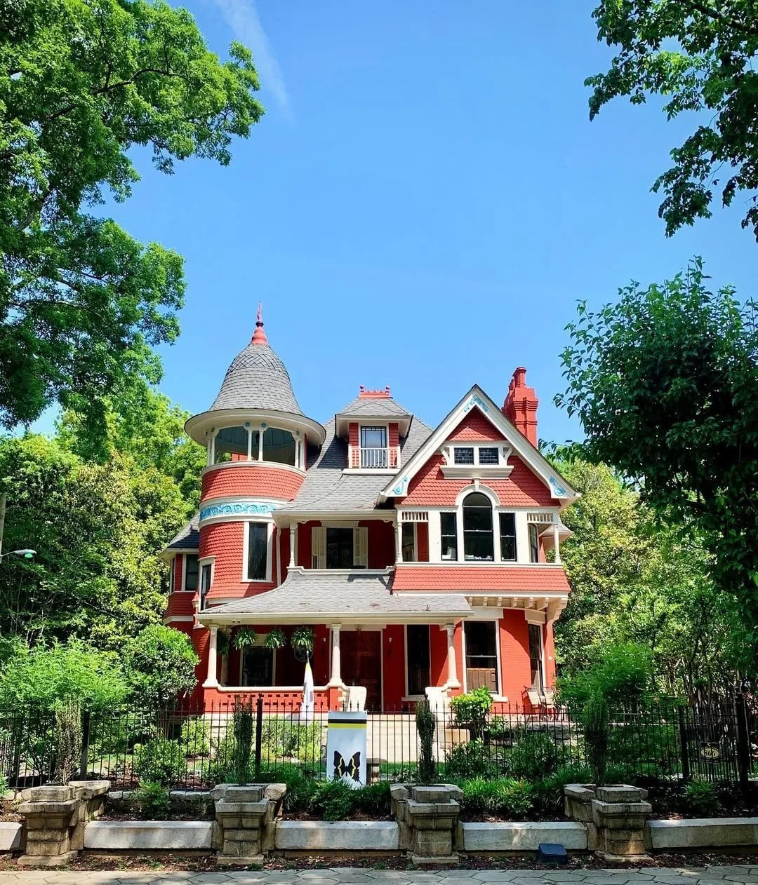 A large Victorian-style house in Inman Park, Atlanta featuring a rounded turret with a conical roof, multiple gables, and a porch with columns, surrounded by green trees and a garden, under a bright blue sky.