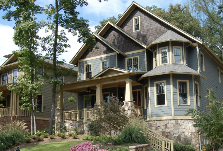 A large, multi-story house with gray siding and a stone foundation, surrounded by trees and landscaped yard.