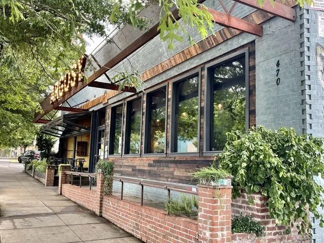 A sidewalk cafe with large window storefront and brick accents, surrounded by greenery, in an urban area.