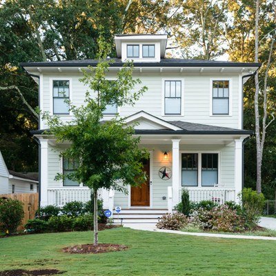 A white three-story house with a front porch, surrounded by a green lawn, trees, and shrubs, under a clear sky.