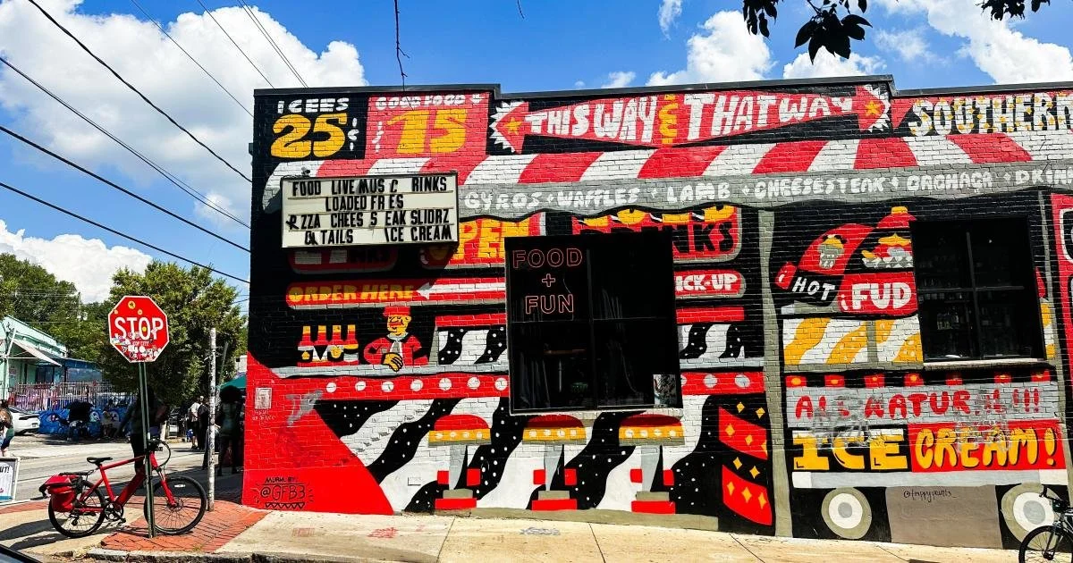 Colorful mural on a building in East Atlanta Village featuring illustrations of burgers, fries, hot dogs, and ice cream, with text promoting food and fun. There is a stop sign and a red bicycle in front of the mural.