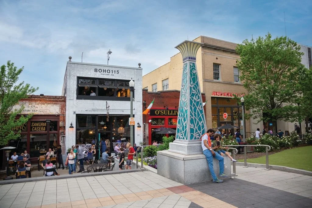 Street view of a lively downtown area with people walking, sitting, and dining outside. Several buildings including a two-story restaurant, a storefront, and ice cream shop, and a decorative blue and cream column with an open courtyard and trees.