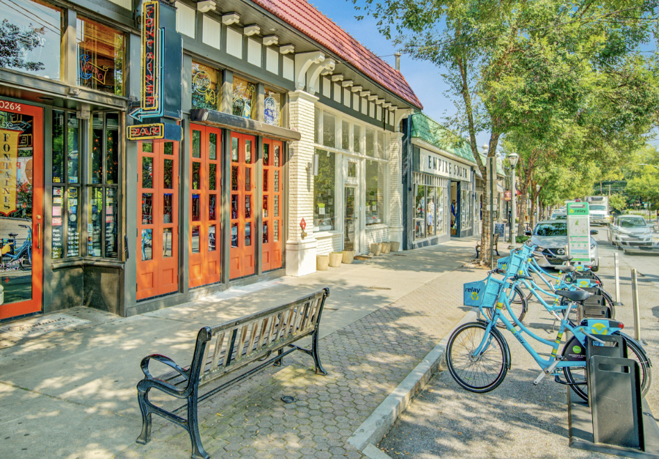 Street scene in Virginia-Highland with storefront shops, a bench, and bikes parked along the curb under trees on a sunny day.