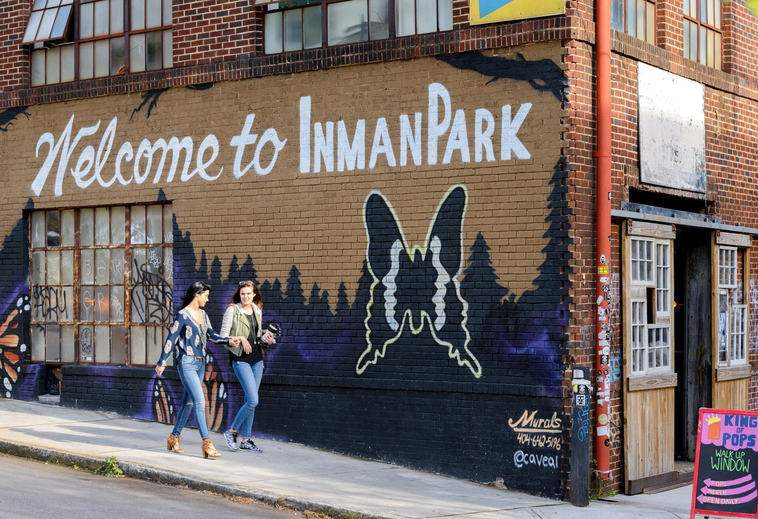 A mural on a brick building in Inman Park, Atlanta with a butterfly and trees painted underneath. Two women walk past the mural, one smiling, carrying drinks.