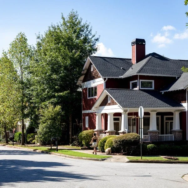 A large red and white house with a front porch and a chimney, surrounded by green trees and bushes, on a residential street with a person walking nearby.