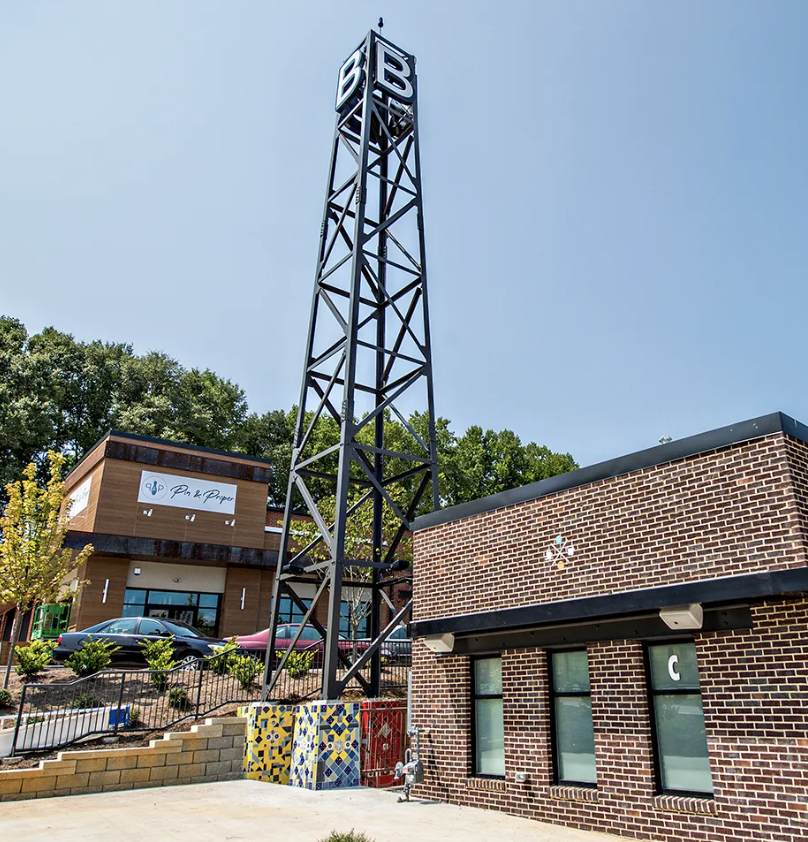 A tall metal tower with the letters B B at the top, next to a brick building with three windows, a fire hydrant, and a colorful mosaic base, with a commercial building and parked cars in the background.