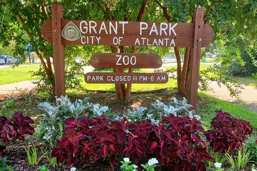 Wooden sign for Grant Park Zoo in Atlanta, with park hours noted, surrounded by colorful flowers and green trees.