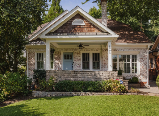 Front view of a brick house with a porch, green grass lawn, and trees in the background.