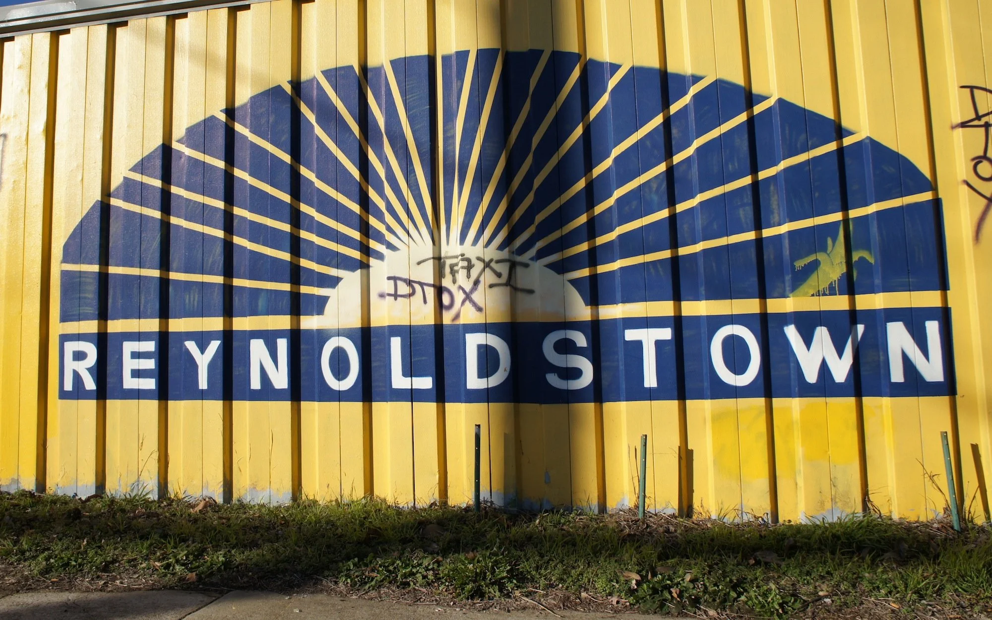 A painted sign on a yellow metal fence with a blue and white sunburst design in Reynoldstown, Atlanta.