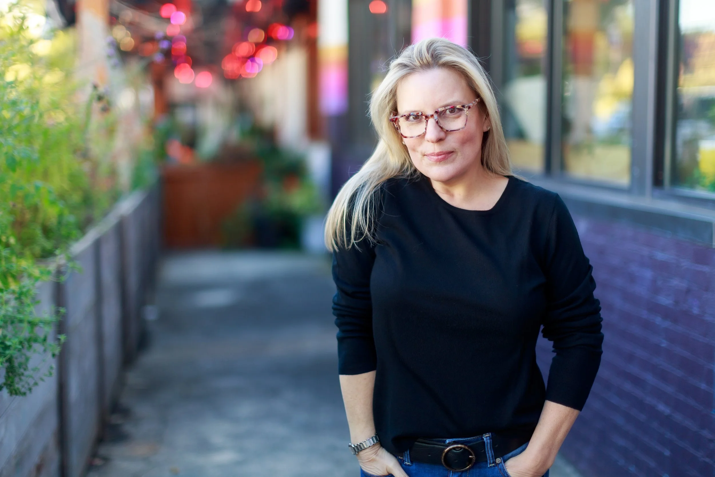 A woman with blonde hair, glasses, and a black shirt standing outdoors near a building with large windows and greenery, during daytime.