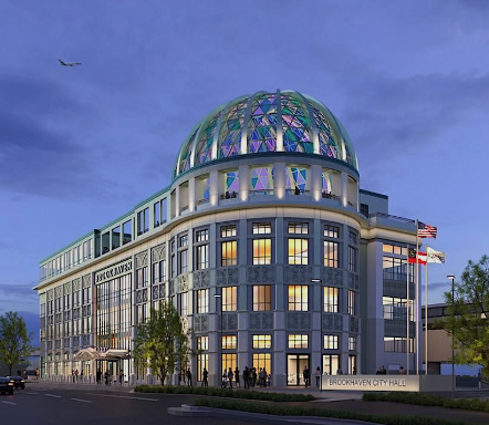 Brookhaven City Hall with a glass dome at dusk, American flags flying outside, people walking, and a clear sky with an airplane overhead.
