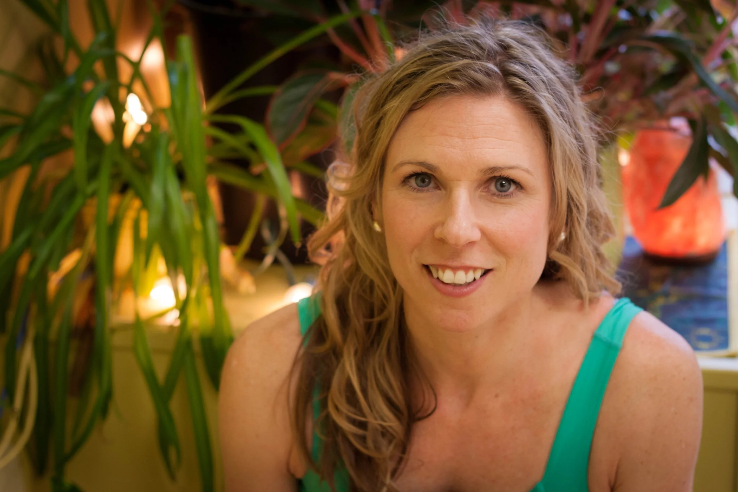 A smiling woman with wavy blonde hair wearing a green tank top, surrounded by green plants and a decorative red clay pot in the background.