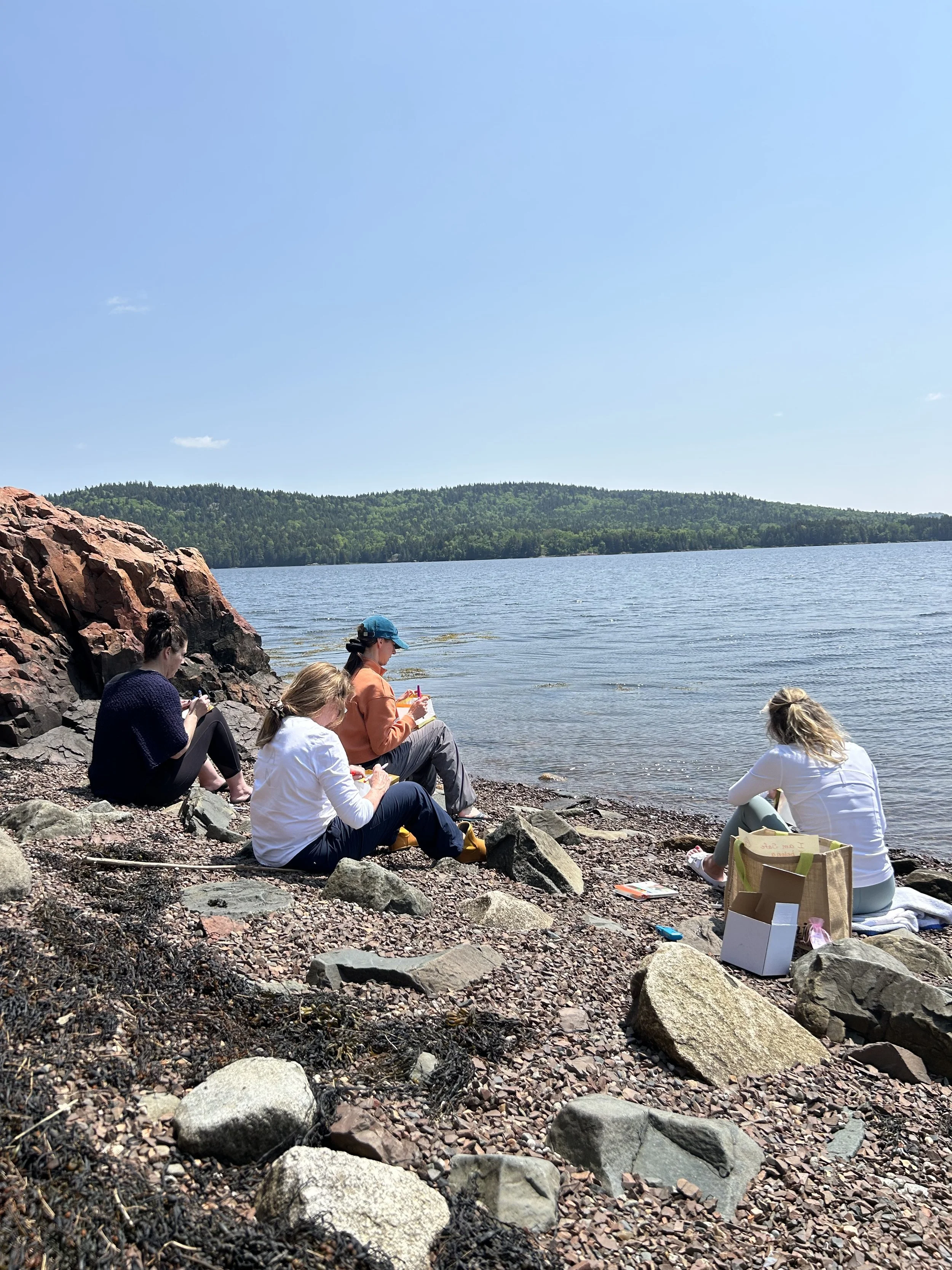 Four women sitting on rocky shoreline near a lake, with trees and hills in the background, enjoying a picnic under a clear blue sky.
