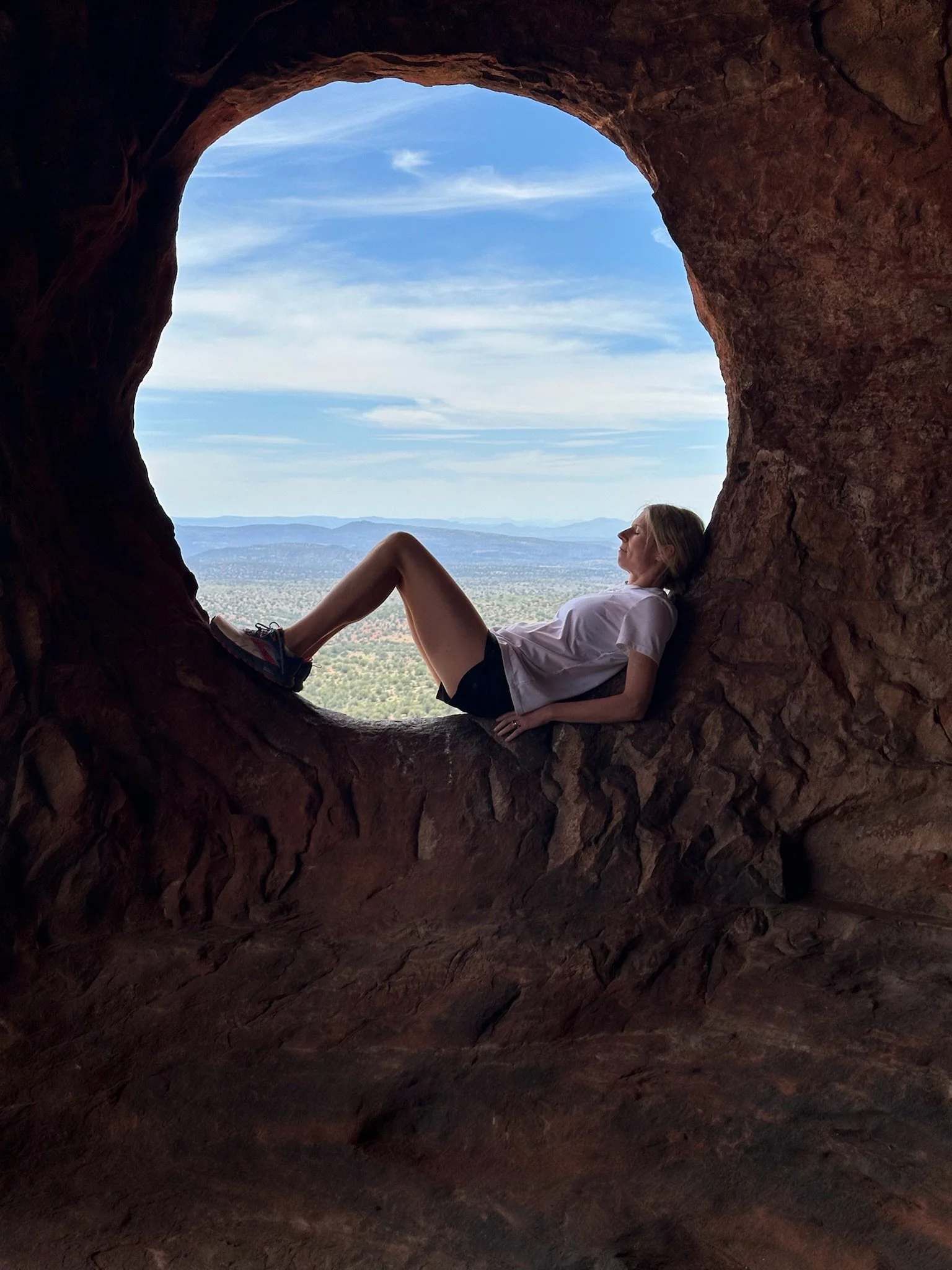 A person relaxing inside a natural rock formation with a circular opening, overlooking a scenic landscape of distant mountains, sky, and greenery.