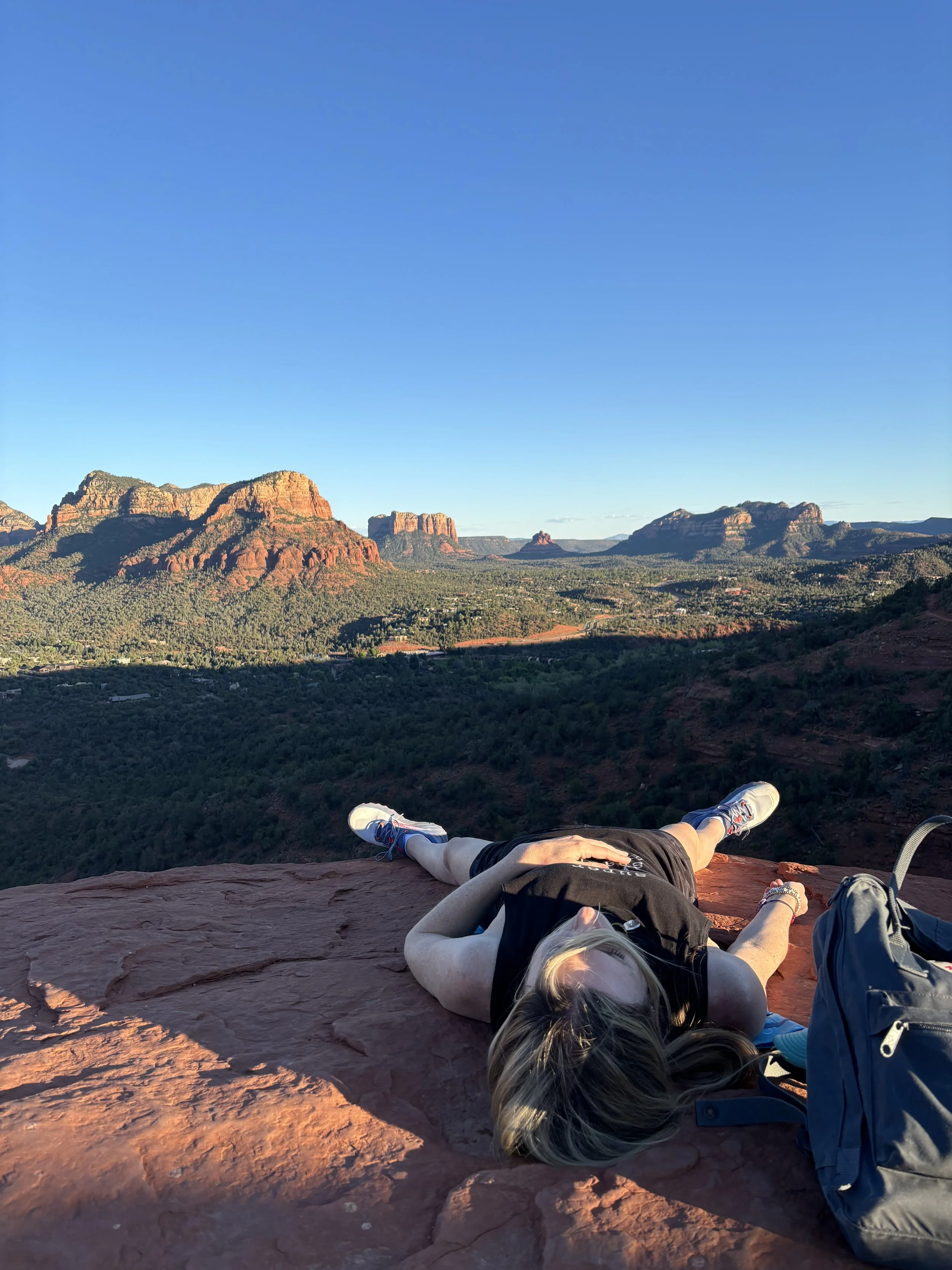 Person lying on a large red rock cliff edge overlooking a landscape of green forests and distant red rock formations under a clear blue sky.