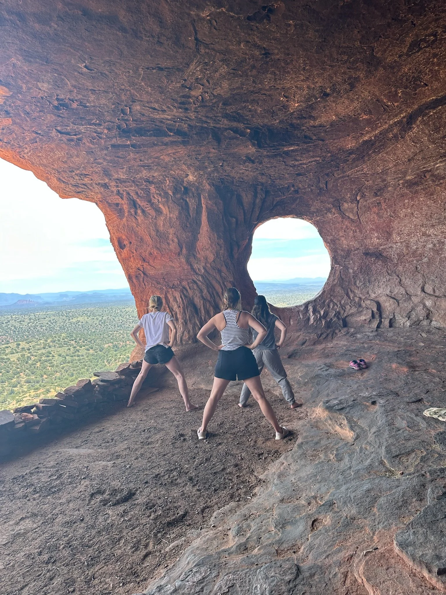 Three women stand in front of a natural rock formation with a large round hole, overlooking a vast desert landscape.