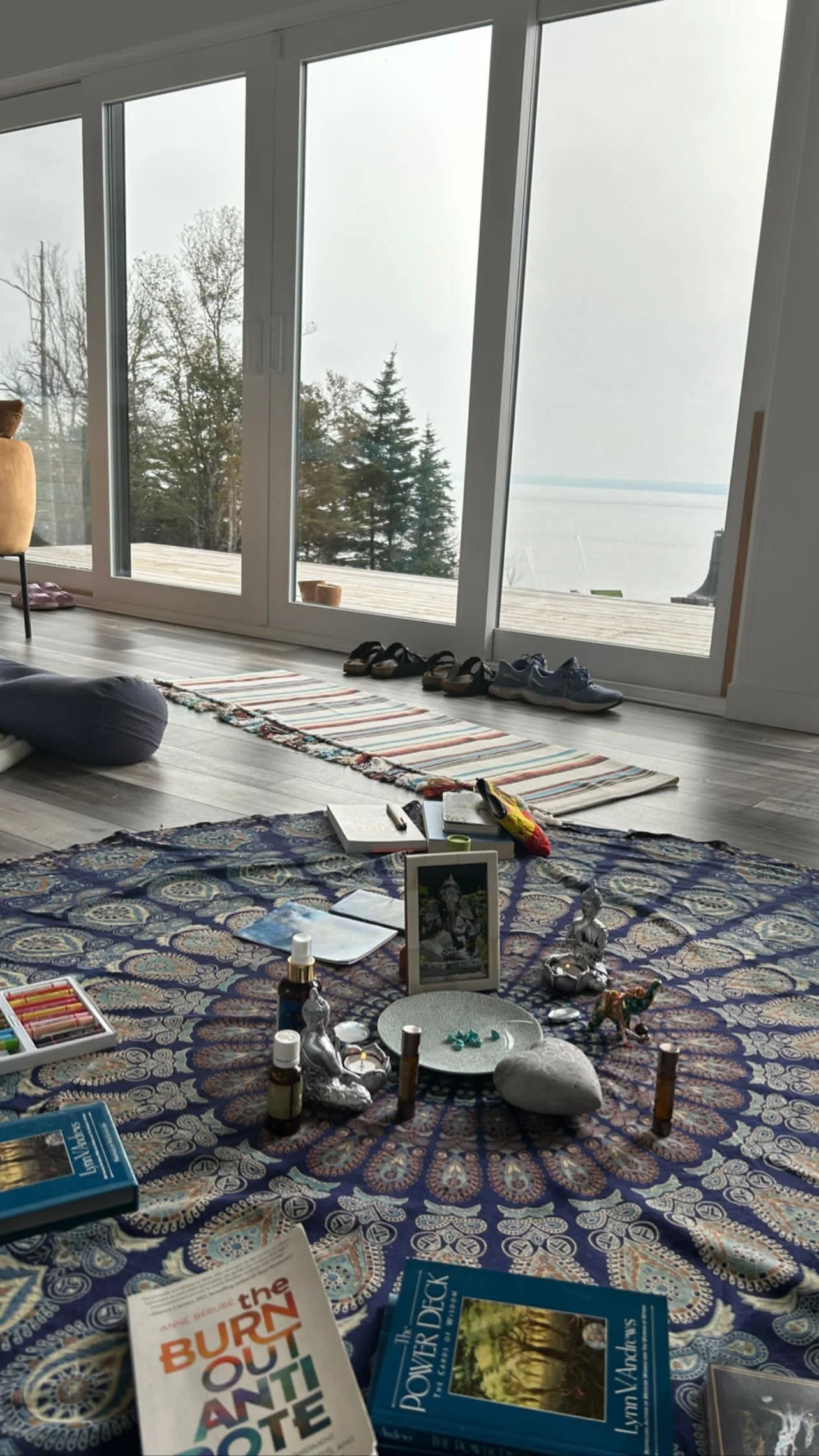 Indoor scene with a view of trees and water through large glass windows, a decorated tapestry on the floor with books, bottles, and spiritual items, and shoes outside the window.