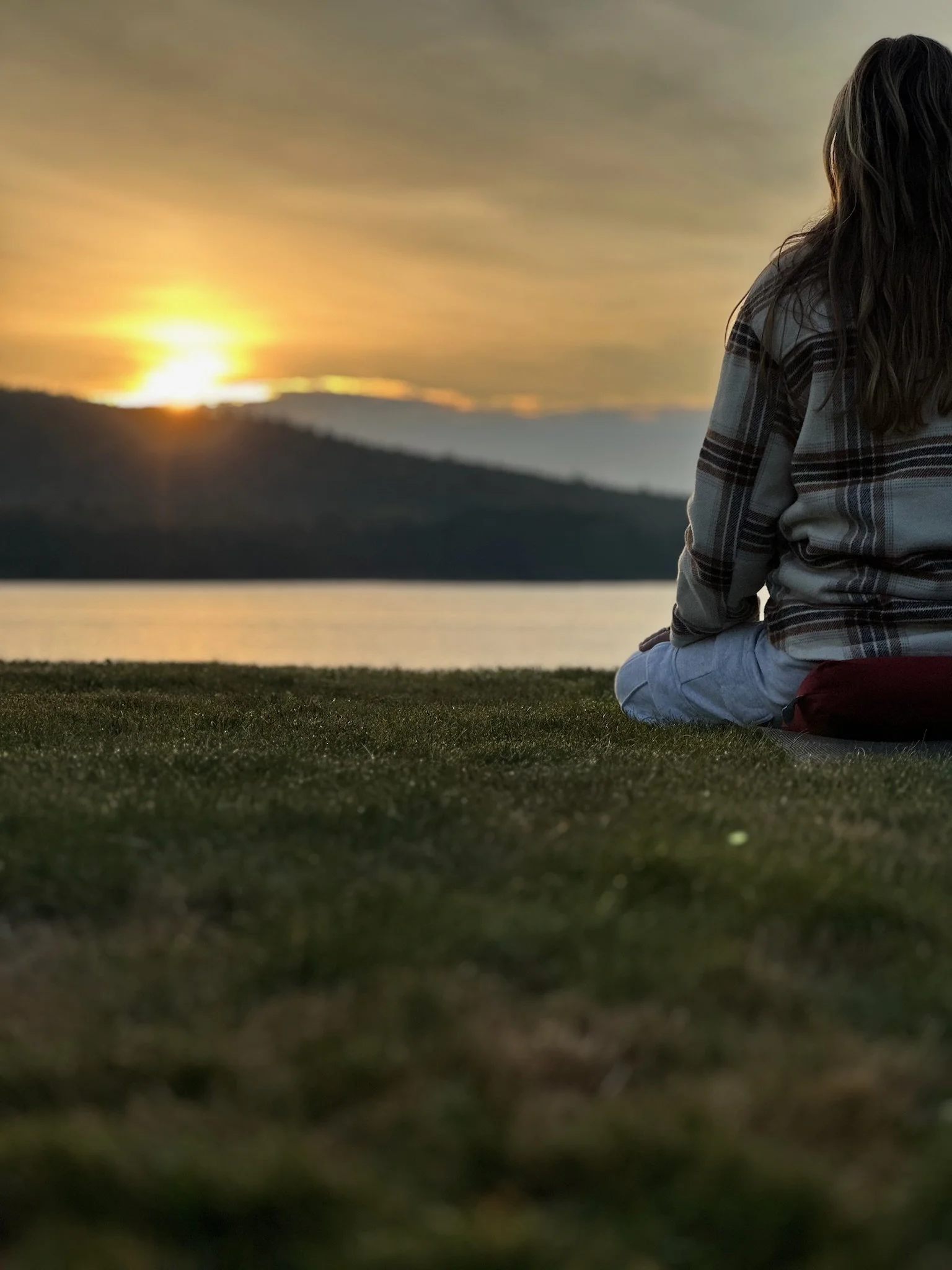 A person sitting on the grass by the water, watching the sunset over distant mountains.