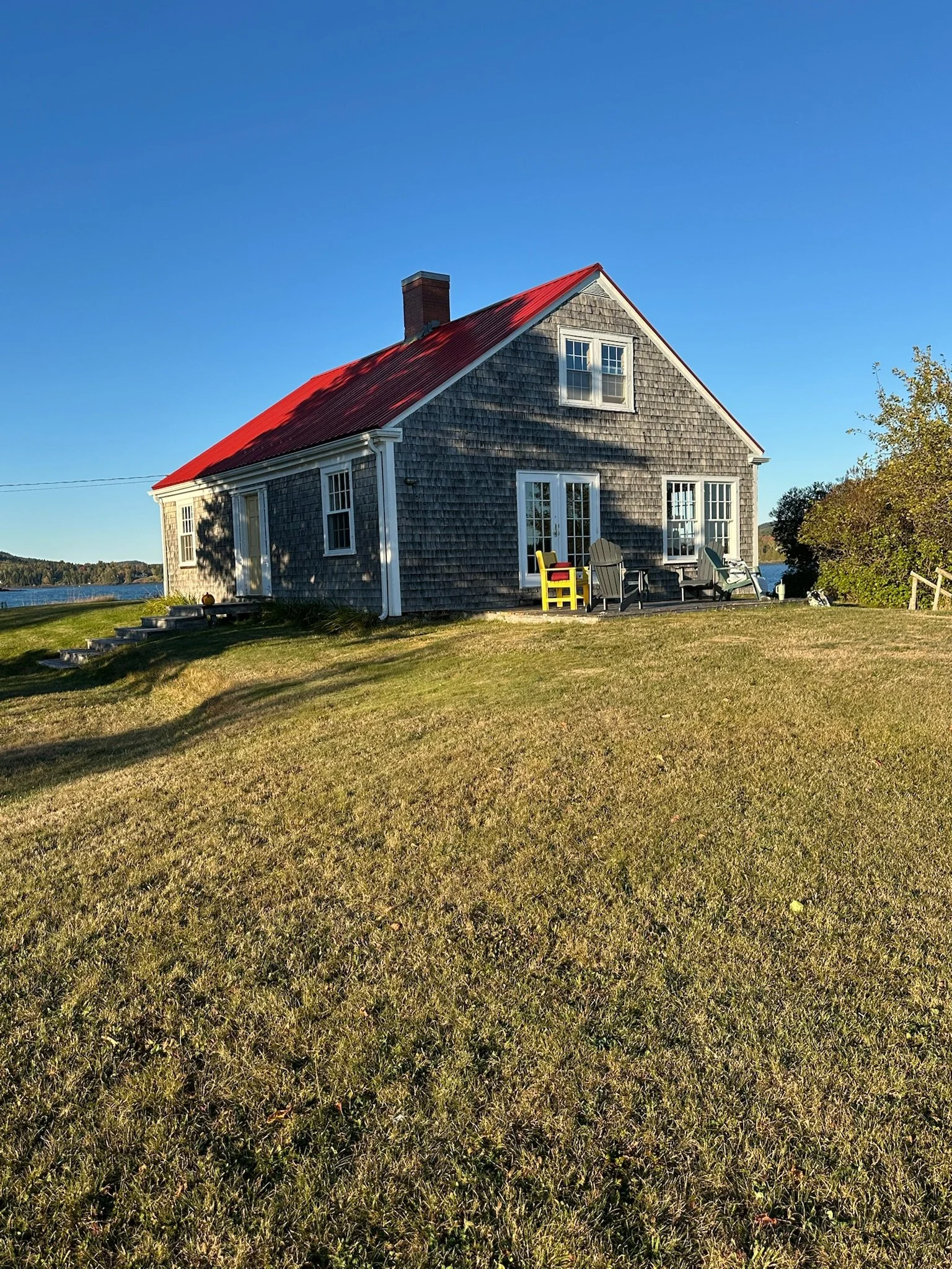 A small, weathered house with gray shingles and a red metal roof, situated on a grassy lawn near a body of water. There are outdoor chairs on the porch and trees around the house, with a clear blue sky overhead.