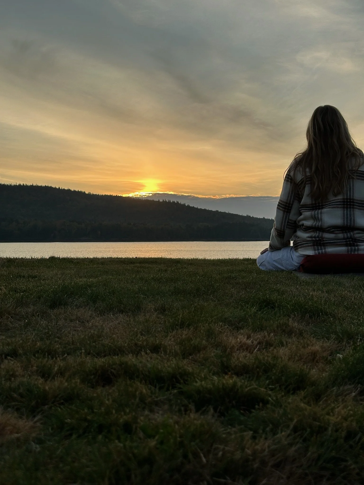 A person with long hair sitting on the grass by a lake, watching the sunset with a mountain range in the background.