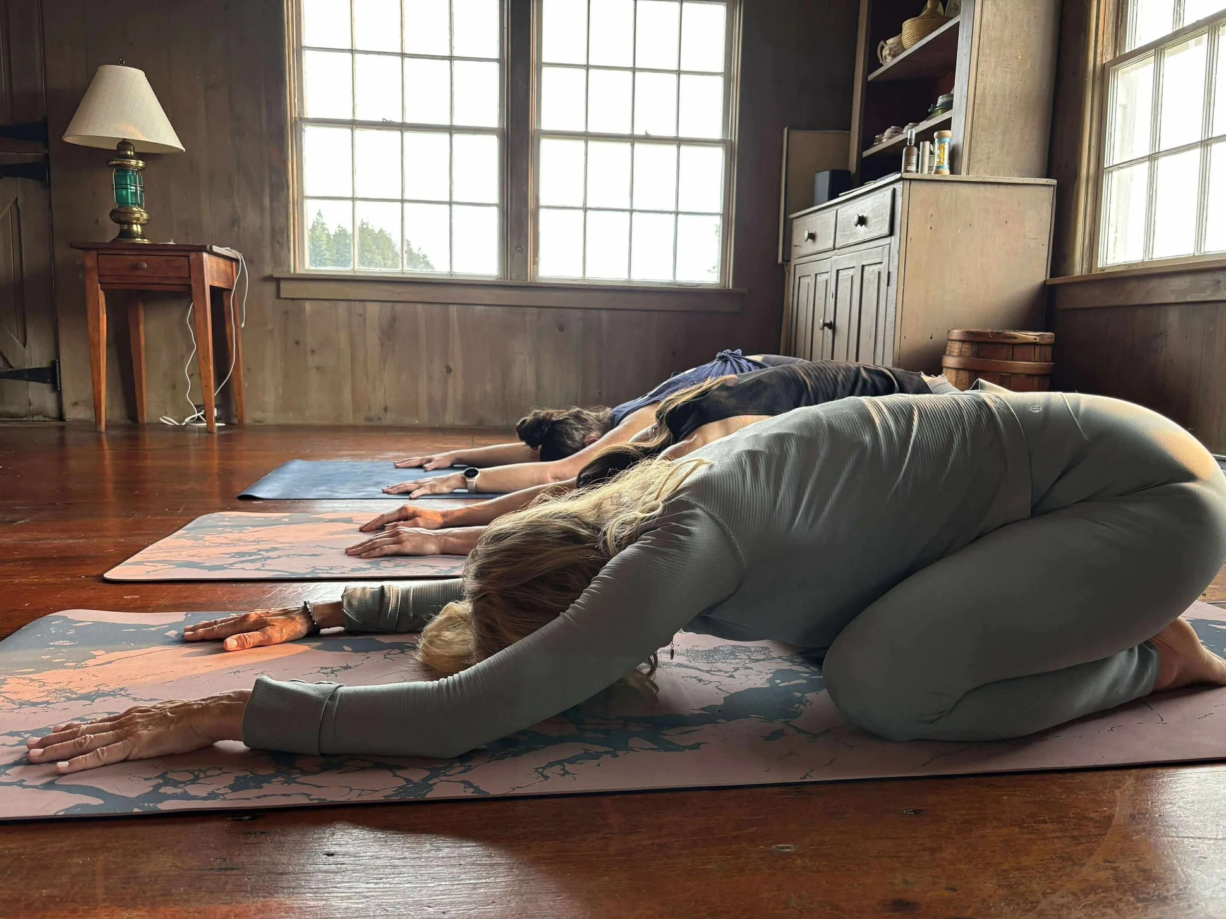 Four people practicing yoga in a wooden-floored room with big windows, wearing workout clothes, in child's pose on yoga mats.