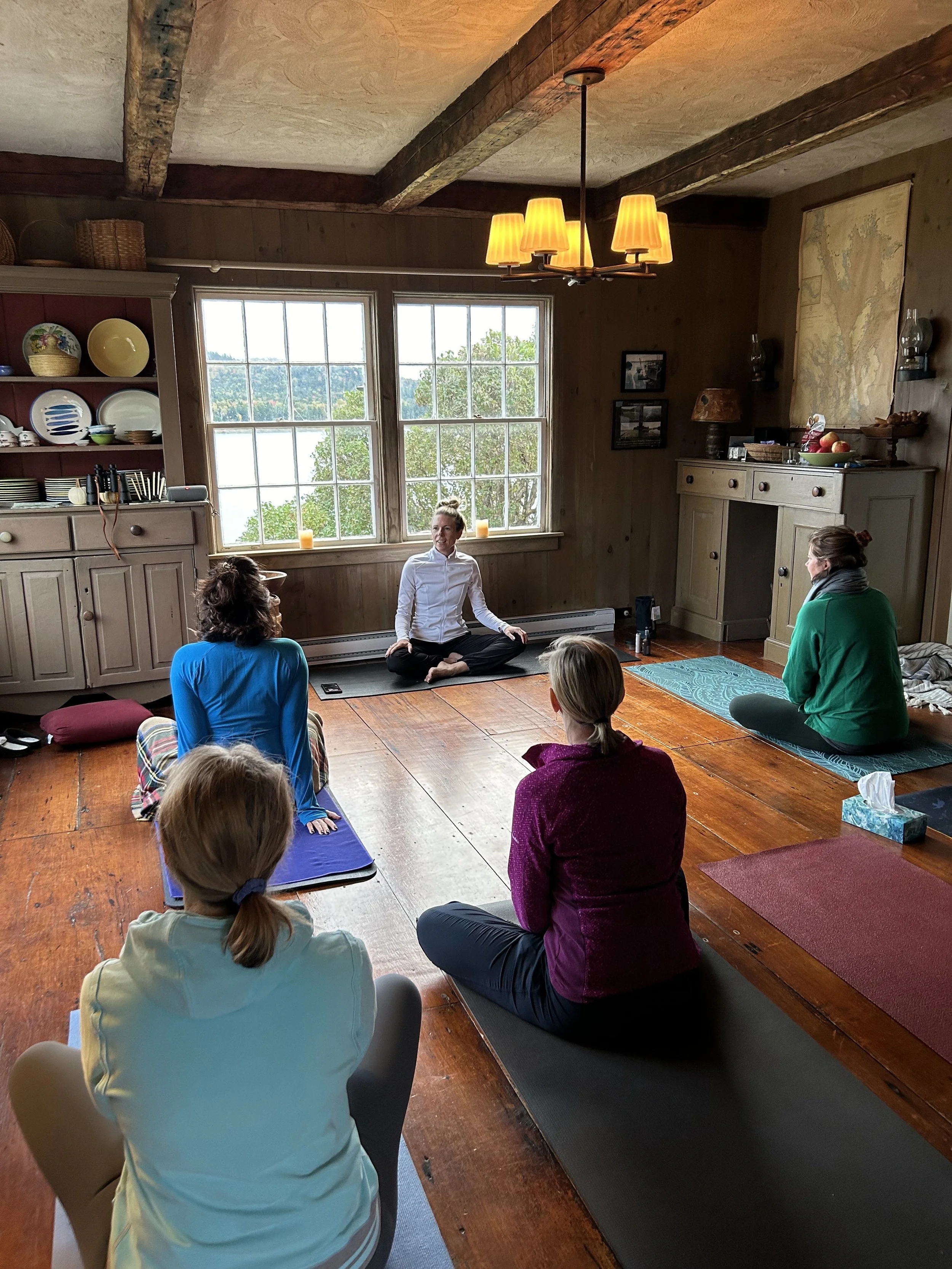 People participating in a yoga or meditation class in a cozy, rustic room with large windows, wooden floors, and a warm lighting fixture.